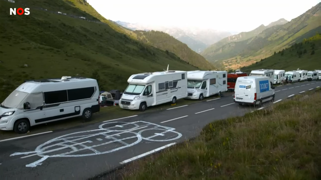Photographe of a large drawing vaguely ressembling an owl, painted on a mountain road in France. If you look closely, you can see that it was originally a penis drawing, which has been covered up. The road is lined with camper vans waiting for the Tour de France racers to arrive.